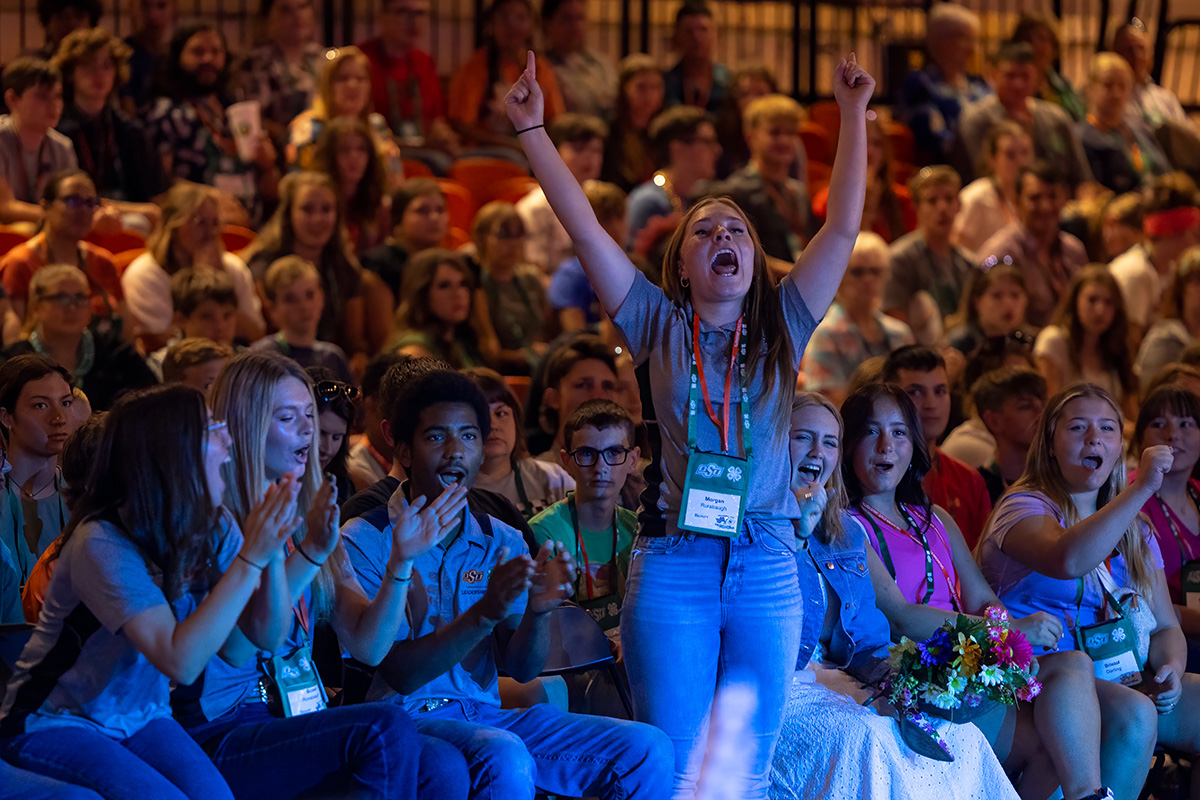 A 4-H member stands up, arms raised in celebration, in front of a crowd at Roundup. A 4-H member stands up, arms raised in celebration, in front of a crowd at Roundup.
